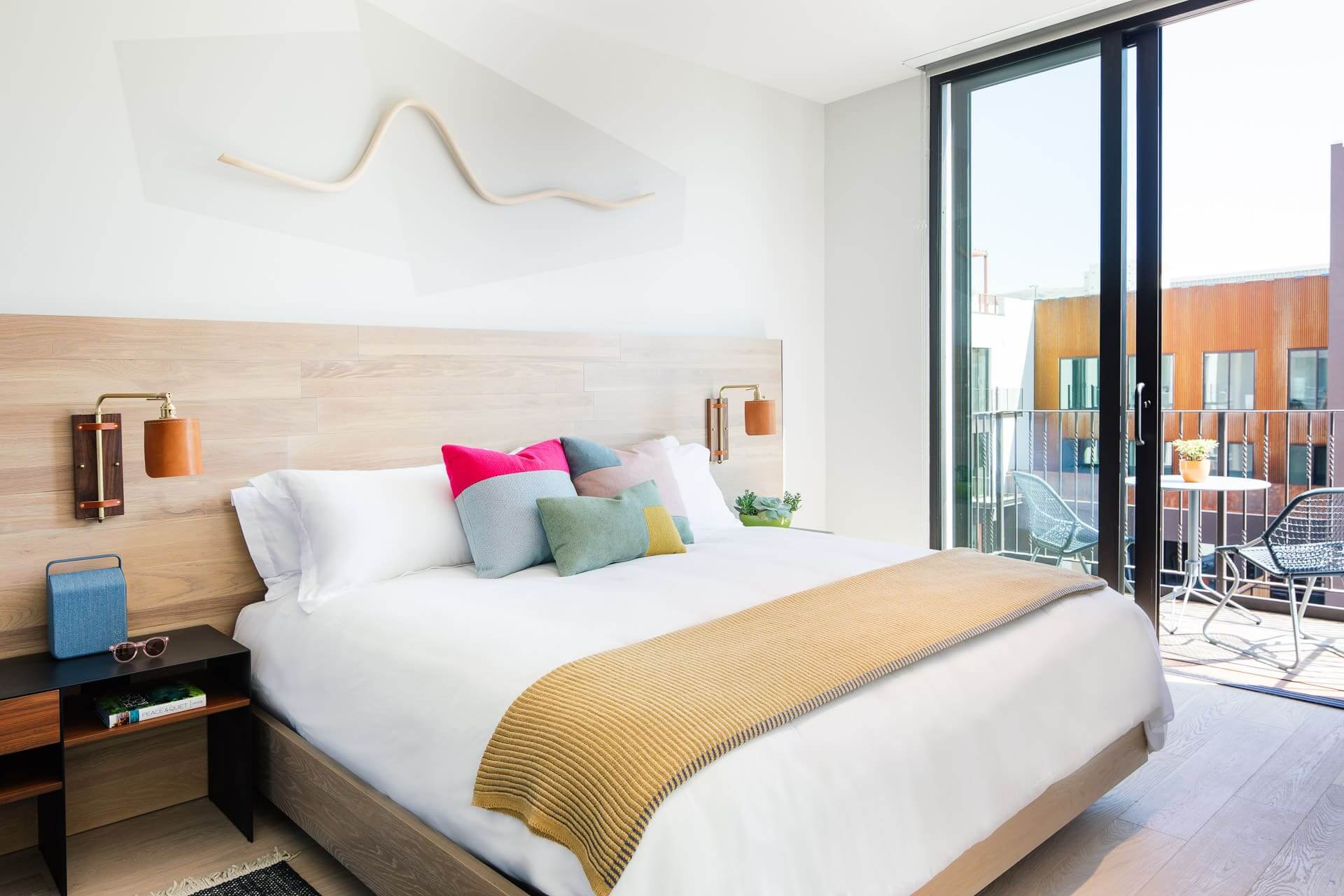 A clean bed with white sheets and colorful pillows, with a view out of a sliding glass door of the Hotel San Luis Obispo room's balcony.